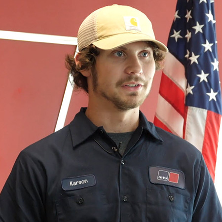 Karson King wearing a baseball cap and dark blue MTU shirt standing in front of red wall and American flag